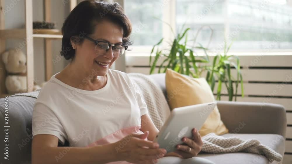 Happy mature woman is making video calling using tablet sitting on couch, beautiful female is holding device, talking, looking at screen, smiling on soft sofa in home atmosphere. Concept Happy mature woman is making video calling using tablet sitting on couch, beautiful female is holding device, talking, looking at screen, smiling on soft sofa in home atmosphere. Concept