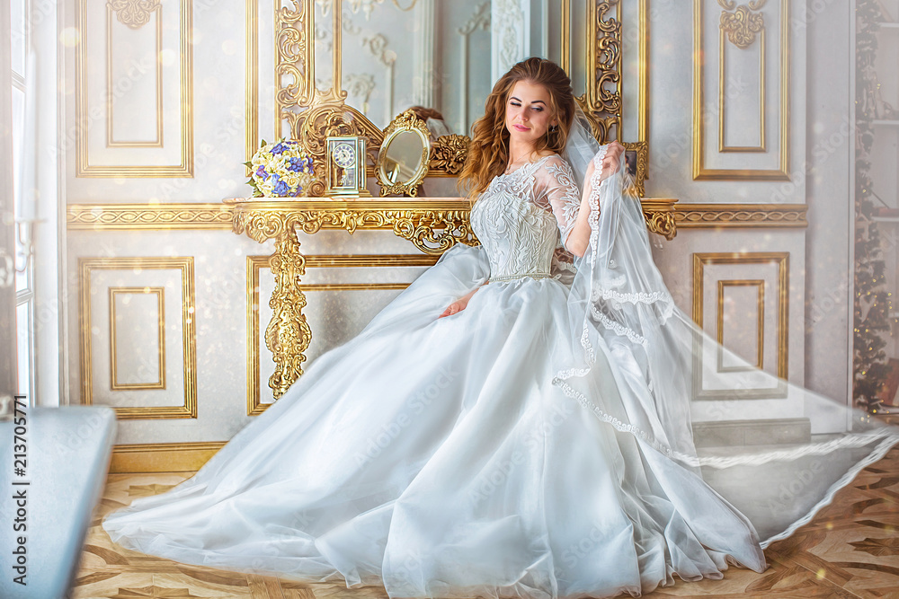 Delightful bride in long wedding dress, sits on chair, in luxurious gold room with large mirror