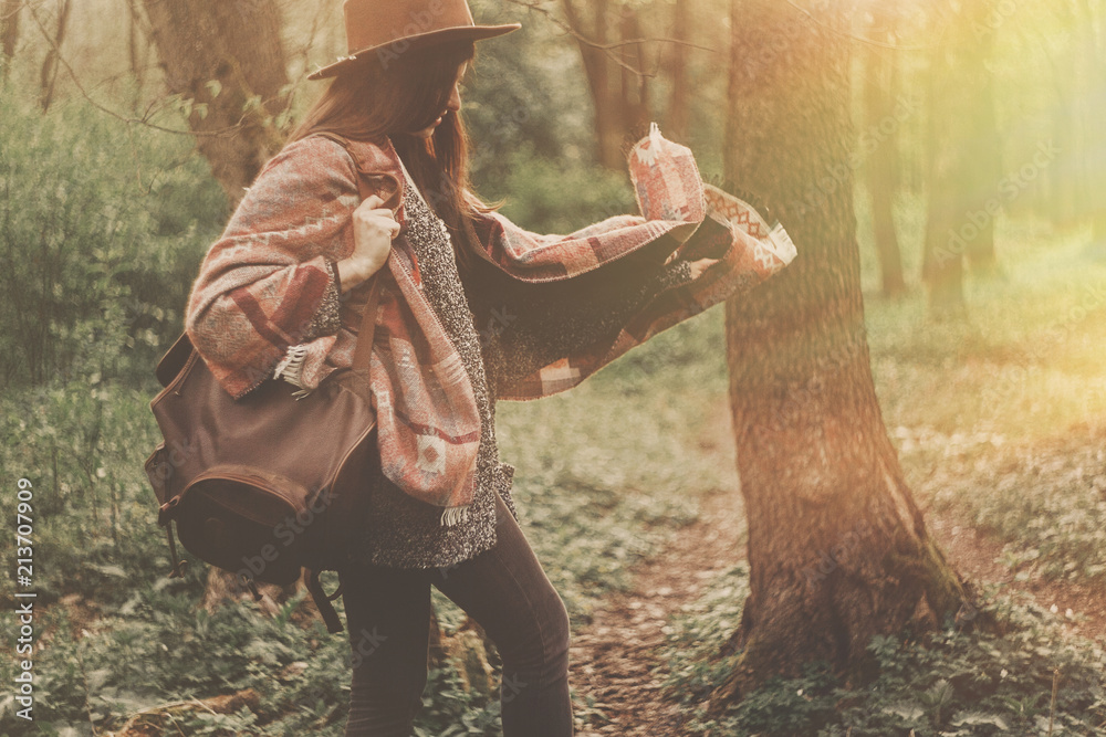stylish hipster traveler girl in hat with backpack exploring in woods in amazing evening sunshine light. woman having fun in sunlight. space for text. atmospheric moment. wanderlust
