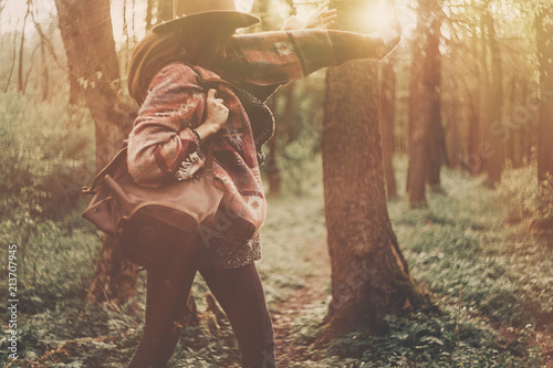 stylish hipster traveler girl in hat with backpack exploring in woods in amazing evening sunshine light. woman having fun jumping in sunlight. space for text. atmospheric moment