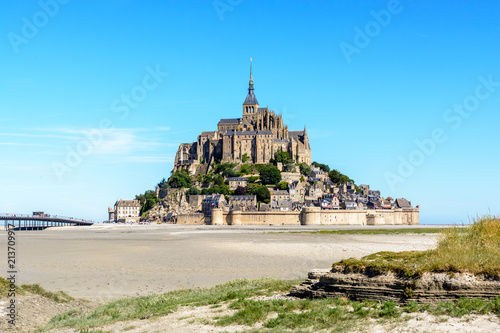 General view of the Mont Saint-Michel tidal island, located in France on the limit between Normandy and Brittany, from the bay at low tide under a summer blue sky.