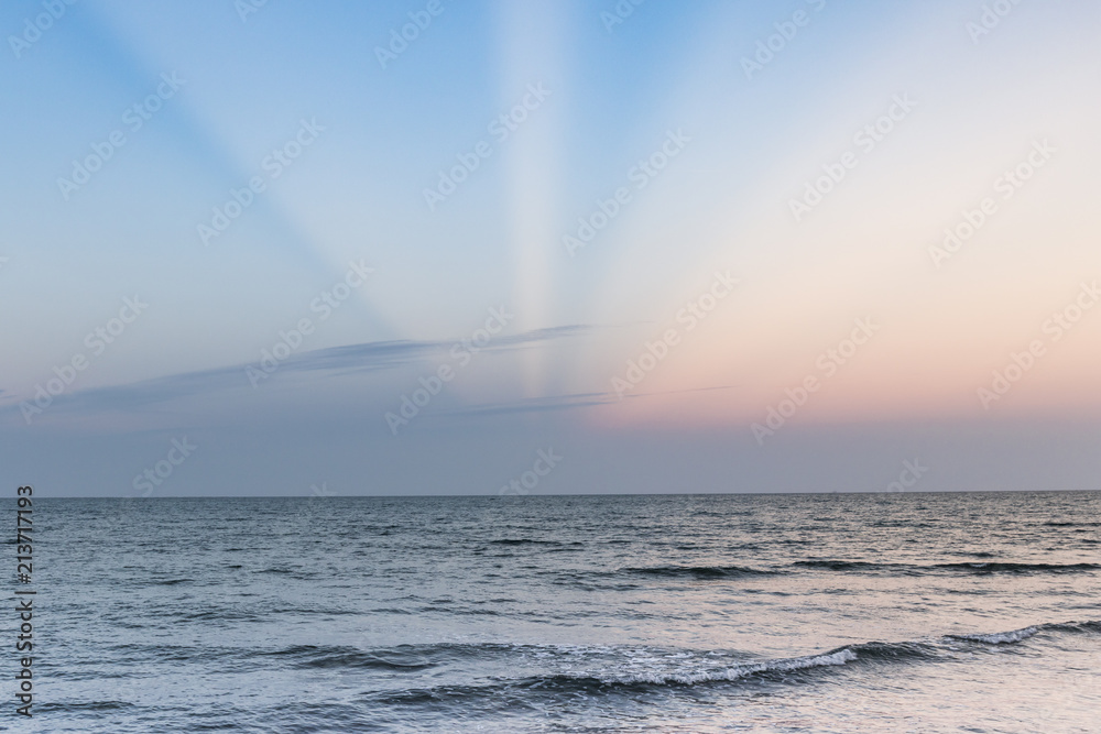 Rayos solares de colores entre nubes en el horizonte de un mar en calma y una pequeña ola. Rayos solares de colores entre nubes en el horizonte de un mar en calma y una pequeña ola.