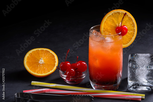 Tequila sunrise cocktail, orange slice, maraschino cherry, straws and ice cube on a dark stone background with space for your text. photo