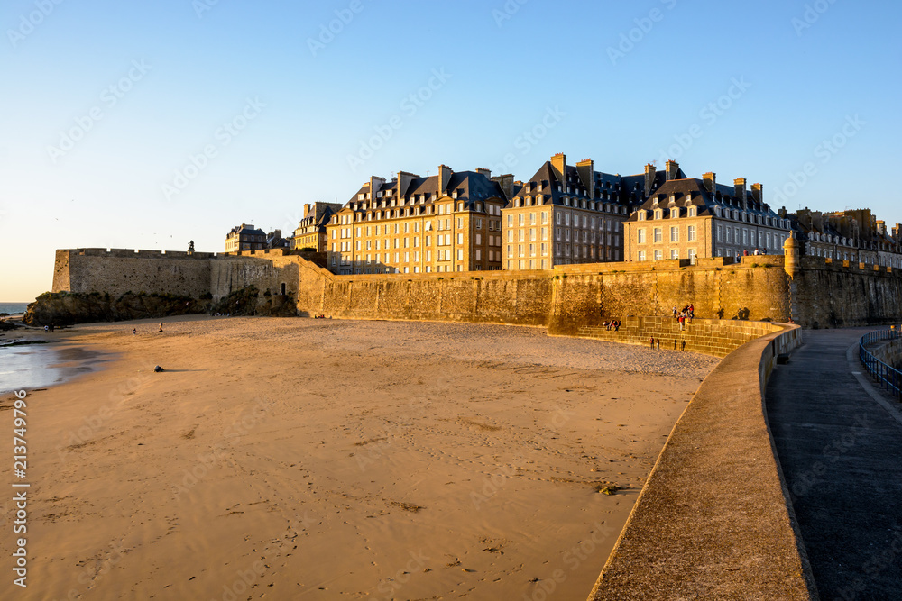 View at sunset of the walled old city of Saint-Malo in Brittany, France, with granite buildings bathed in warm light sticking out above the wall and the Mole beach at the foot of the rampart.