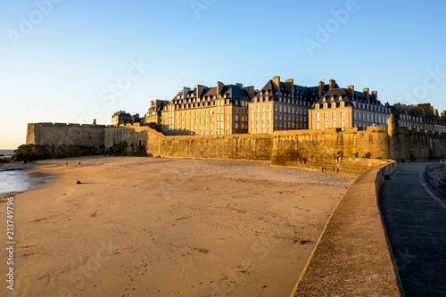 View at sunset of the walled old city of Saint-Malo in Brittany, France, with granite buildings bathed in warm light sticking out above the wall and the Mole beach at the foot of the rampart.
