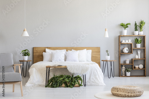 Patterned armchair and pouf in bright bedroom interior with wooden bed and plants. Real photo