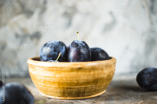 Ripe blue plums in a clay bowl on a gray table. Summer seasonal fruit concept