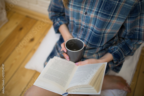 Beautiful girl is sitting on the wooden floor. Young woman in men's checkered shirt is drinking coffee and reading a book on white pillow. Cozy morning with a mug in hands.