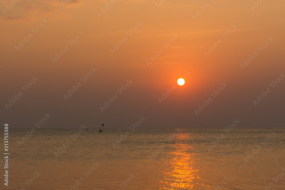 Person practicing windsurfing on the distance at warm sunset reflected on the calm sea in the island of Koh Phangan, Thailand. Summer holidays leisure activity, perfect vacation concepts