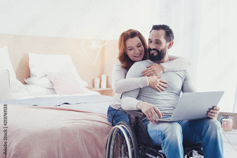 Pleasant morning. Young emotional man sitting in a wheelchair with a laptop in his hands while his kind attentive wife hugging him Pleasant morning. Young emotional man sitting in a wheelchair with a laptop in his hands while his kind attentive wife hugging him