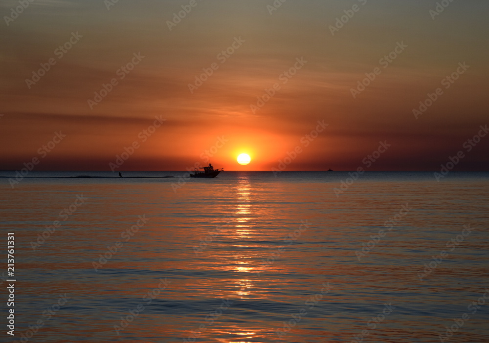 A cruising sail boat passes majestically beneath the enchanting sunset. The Sun casts orange shades across an evening sky at Mindil Beach (Darwin, Northern Territory, Australia).