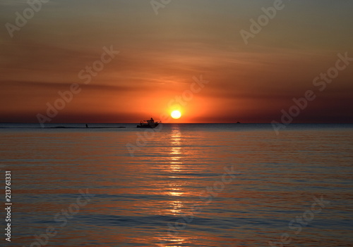 A cruising sail boat passes majestically beneath the enchanting sunset. The Sun casts orange shades across an evening sky at Mindil Beach (Darwin, Northern Territory, Australia).