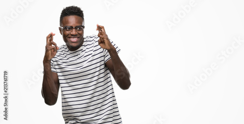 Young african american man wearing glasses and navy t-shirt smiling crossing fingers with hope and eyes closed. Luck and superstitious concept.