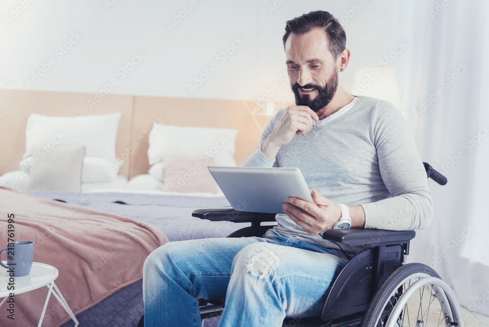 Interesting idea. Young creative ambitious blogger sitting in a wheelchair and smiling while looking at the screen of his modern device and getting an interesting idea