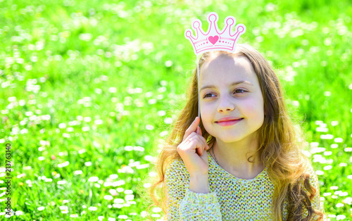 Girl on dreamy face spend leisure outdoors. Princess concept. Child posing with cardboard crown for photo session at meadow. Girl sits on grass at grassplot  green background.