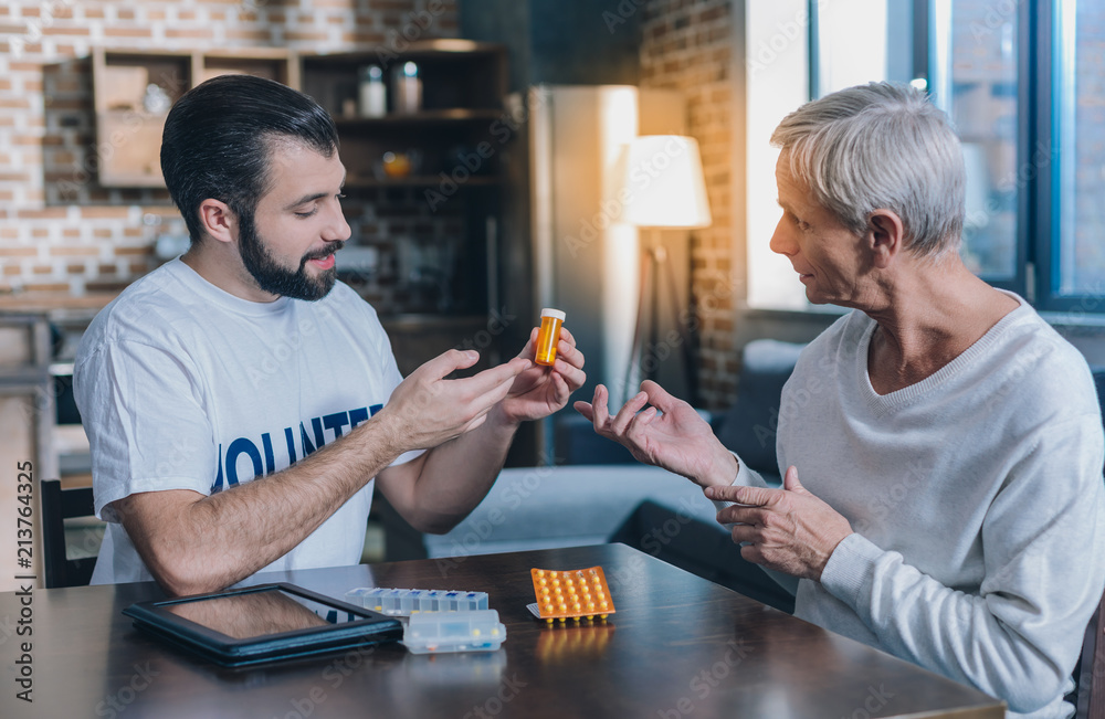 Good to heath. Attractive content young man smiling and holding some pills while talking with an old man Good to heath. Attractive content young man smiling and holding some pills while talking with an old man