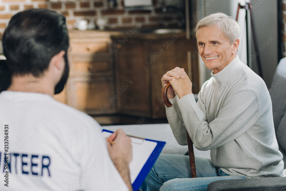 I feel happy. Inspired kind grey-haired man smiling and sitting with his stick while talking with a volunteer I feel happy. Inspired kind grey-haired man smiling and sitting with his stick while talking with a volunteer