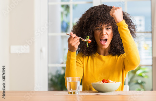 African american woman eating pasta salad at home annoyed and frustrated shouting with anger, crazy and yelling with raised hand, anger concept