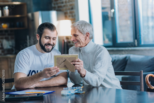 My family. Happy grey-haired man smiling and showing photos to a young man