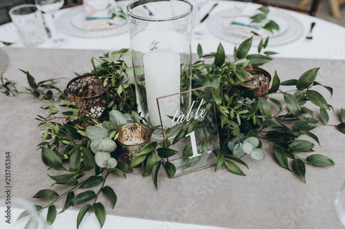 on table with gray tablecloth is a candle in candlestick, plate with number and composition of greenery