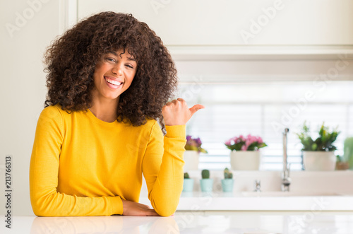 African american woman wearing yellow sweater at kitchen smiling with happy face looking and pointing to the side with thumb up.