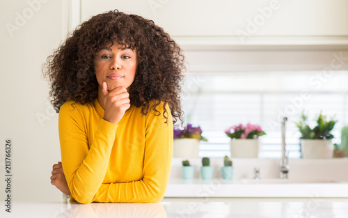 African american woman wearing yellow sweater at kitchen looking confident at the camera with smile with crossed arms and hand raised on chin. Thinking positive.