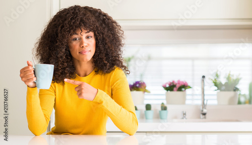 Beautiful african american woman holding a cup of coffee at home very happy pointing with hand and finger