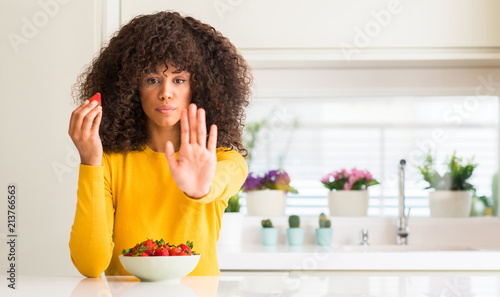African american woman eating strawberries at home with open hand doing stop sign with serious and confident expression  defense gesture