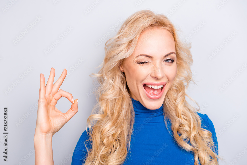 Portrait of sexy, cheerful, stylish, pretty, cute, charming, nice, blonde woman in blue turtleneck showing ok sign with fingers blinking with one eye, open mouth isolated on grey background Portrait of sexy, cheerful, stylish, pretty, cute, charming, nice, blonde woman in blue turtleneck showing ok sign with fingers blinking with one eye, open mouth isolated on grey background