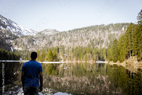 young man with cap and blue shirt is standing in front of mountain lake with raising arms