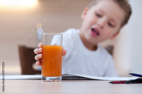 Little cute boy in white T-shirt paint the coloring book pencil at kitchen table next to glass of freshly squeezed juice