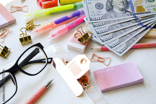 Educations costs concept. Stack of multiple hundred dollar bills, empty check box planner, colorful pens, school supplies, hipster eyeglasses w/ black frame. Background, flat lay, close up, copy space