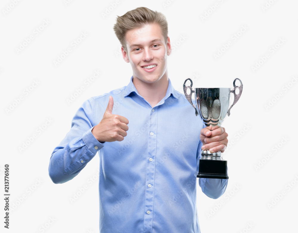 Young handsome blond man holding a trophy happy with big smile doing ok sign, thumb up with fingers, excellent sign Young handsome blond man holding a trophy happy with big smile doing ok sign, thumb up with fingers, excellent sign