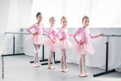 cute multiethnic kids in pink tutu skirts exercising and smiling at camera in ballet studio