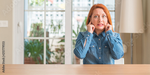 Redhead woman at home covering ears with fingers with annoyed expression for the noise of loud music. Deaf concept.