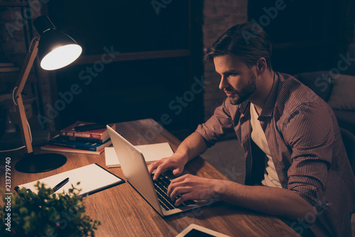 Portrait of attractive programmer, hard worker, busy man in shirt with hairstyle working at night, taking work at home, looking at screen of laptop, sitting in work place, holding hands on keypad photo