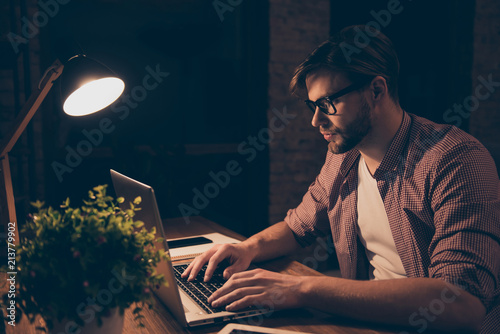 Portrait of attractive programmer, hard worker, busy man in shirt with hairstyle working at night, taking work at home, looking at screen of laptop, sitting in work station, holding hands on keypad photo