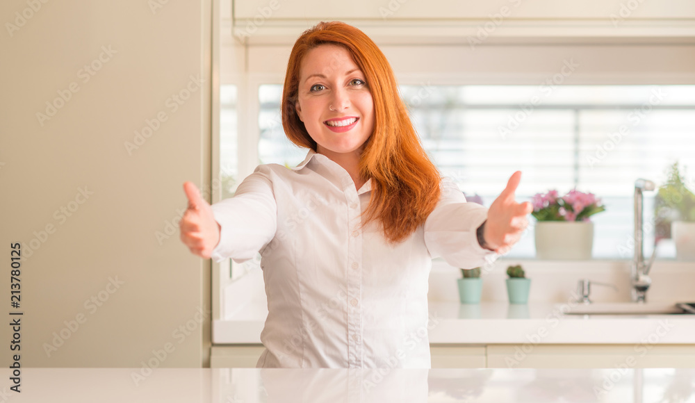 Redhead woman at kitchen looking at the camera smiling with open arms for hug. Cheerful expression embracing happiness.