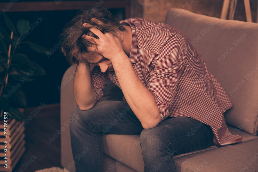 Obraz premium Frustrated young guy with bristle is holding his head, he is upset because his business has failed, sitting on the couch alone, bad luck, relationship problems, suffering from headache