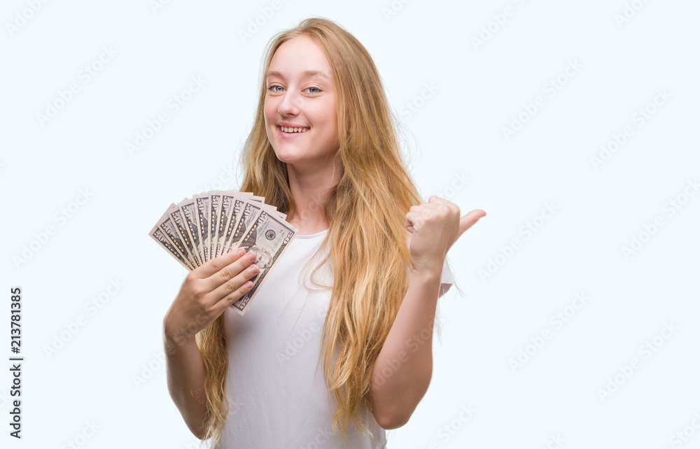 Blonde teenager woman holding dollars pointing and showing with thumb up to the side with happy face smiling Blonde teenager woman holding dollars pointing and showing with thumb up to the side with happy face smiling