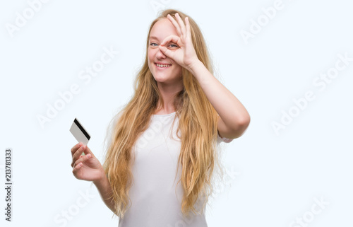 Blonde teenager woman holding credit card with happy face smiling doing ok sign with hand on eye looking through fingers