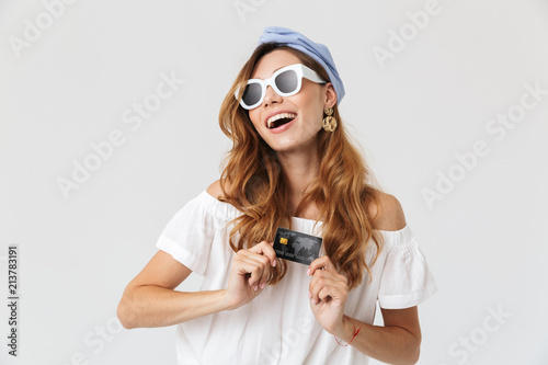 Image of happy young woman 20s wearing sunglasses looking aside and showing credit card, isolated over white background