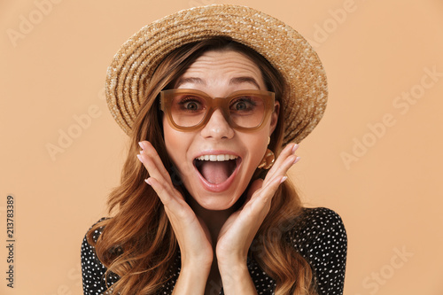 Portrait of adorable woman 20s wearing straw hat and sunglasses posing on camera with happy smile, isolated over beige background