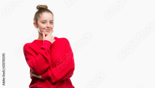 Young blonde woman wearing bun and red sweater looking confident at the camera with smile with crossed arms and hand raised on chin. Thinking positive.