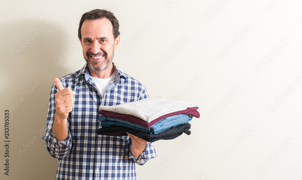 Senior man holding folded laundry clothes happy with big smile doing ok sign, thumb up with fingers, excellent sign Senior man holding folded laundry clothes happy with big smile doing ok sign, thumb up with fingers, excellent sign