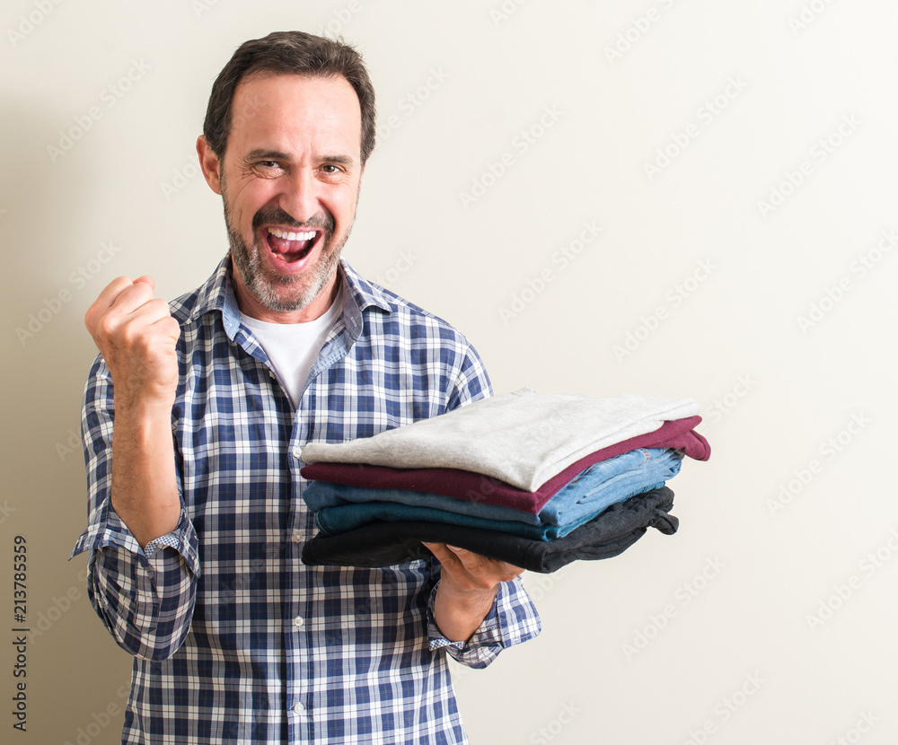 Senior man holding folded laundry clothes screaming proud and celebrating victory and success very excited, cheering emotion Senior man holding folded laundry clothes screaming proud and celebrating victory and success very excited, cheering emotion