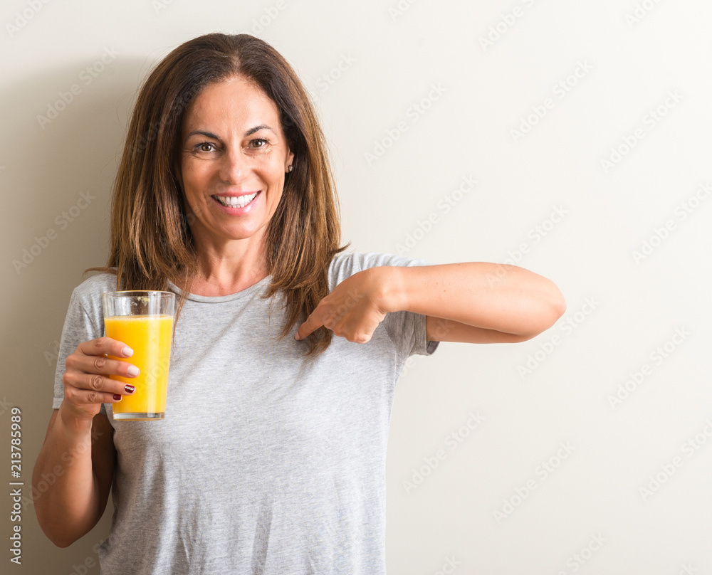 Middle age woman drinking orange juice in a glass with surprise face pointing finger to himself Middle age woman drinking orange juice in a glass with surprise face pointing finger to himself