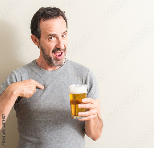Senior man drinking beer with surprise face pointing finger to himself