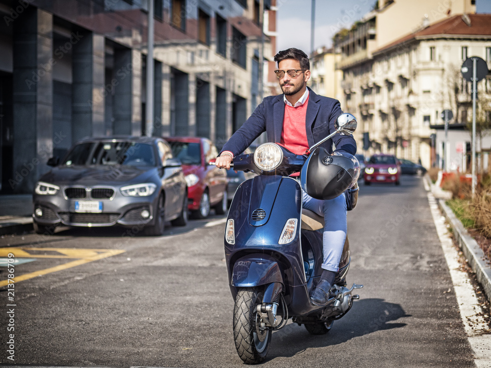 Young stylish man in suit and sunglasses driving scooter on empty road in sunny city.
