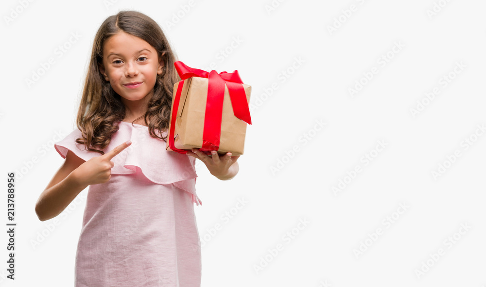 Brunette hispanic girl holding a gift very happy pointing with hand and finger Brunette hispanic girl holding a gift very happy pointing with hand and finger